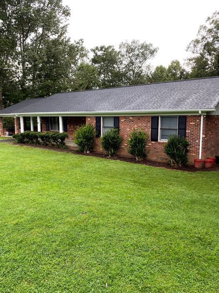 Brick ranch-style house with green lawn and landscaping. Dark roof, white trim, black shutters. Cloudy sky.
