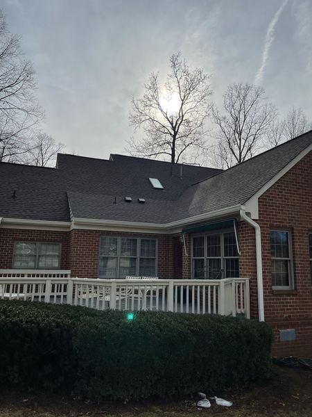 Brick house with a dark roof and a white deck. The sun shines behind bare trees.