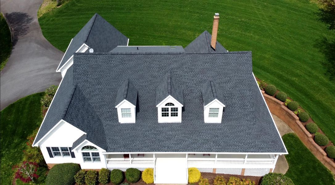 Dark gray shingled roof on a white house with three dormers, a chimney, and green grass.