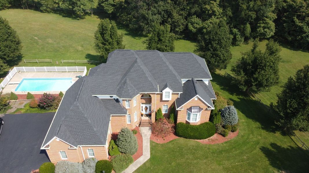 Aerial view of a large two-story brick house with a dark gray roof, a pool, and surrounding green lawn and trees.