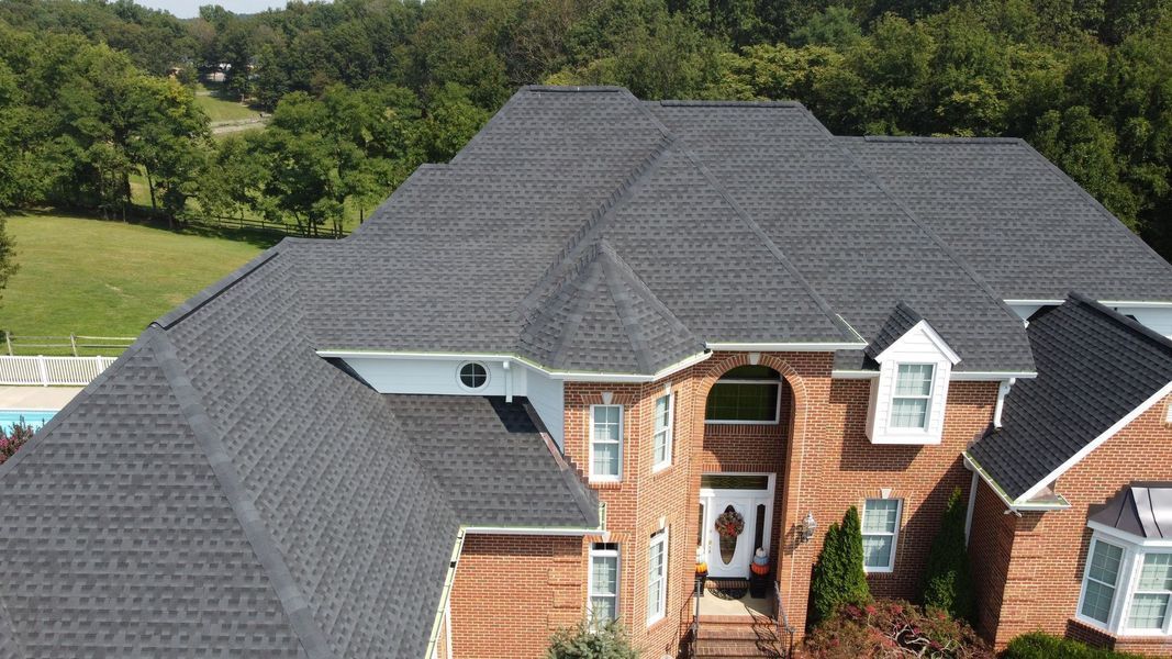 Brick house with dark gray shingle roof, surrounded by trees and a glimpse of a pool.