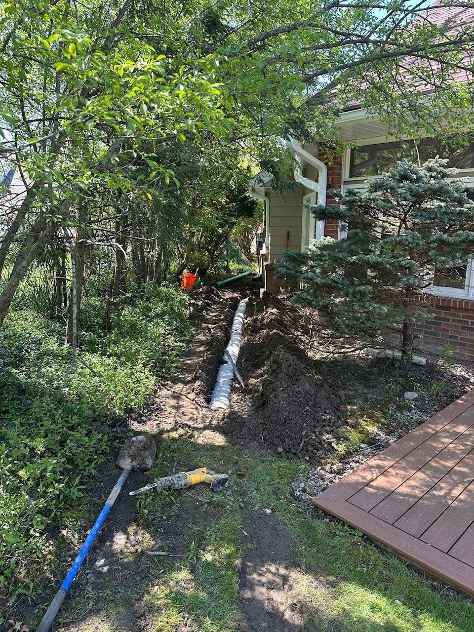 A trench dug alongside a house, with a pipe installed, likely for drainage. Dirt, tools, and greenery surround the construction.