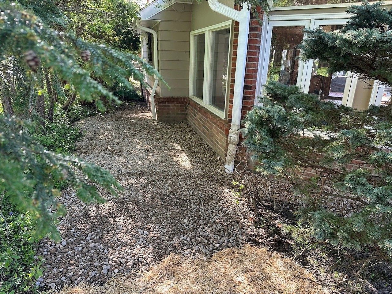 A gravel-covered area beside a beige house with brick foundation, framed by green bushes and trees.