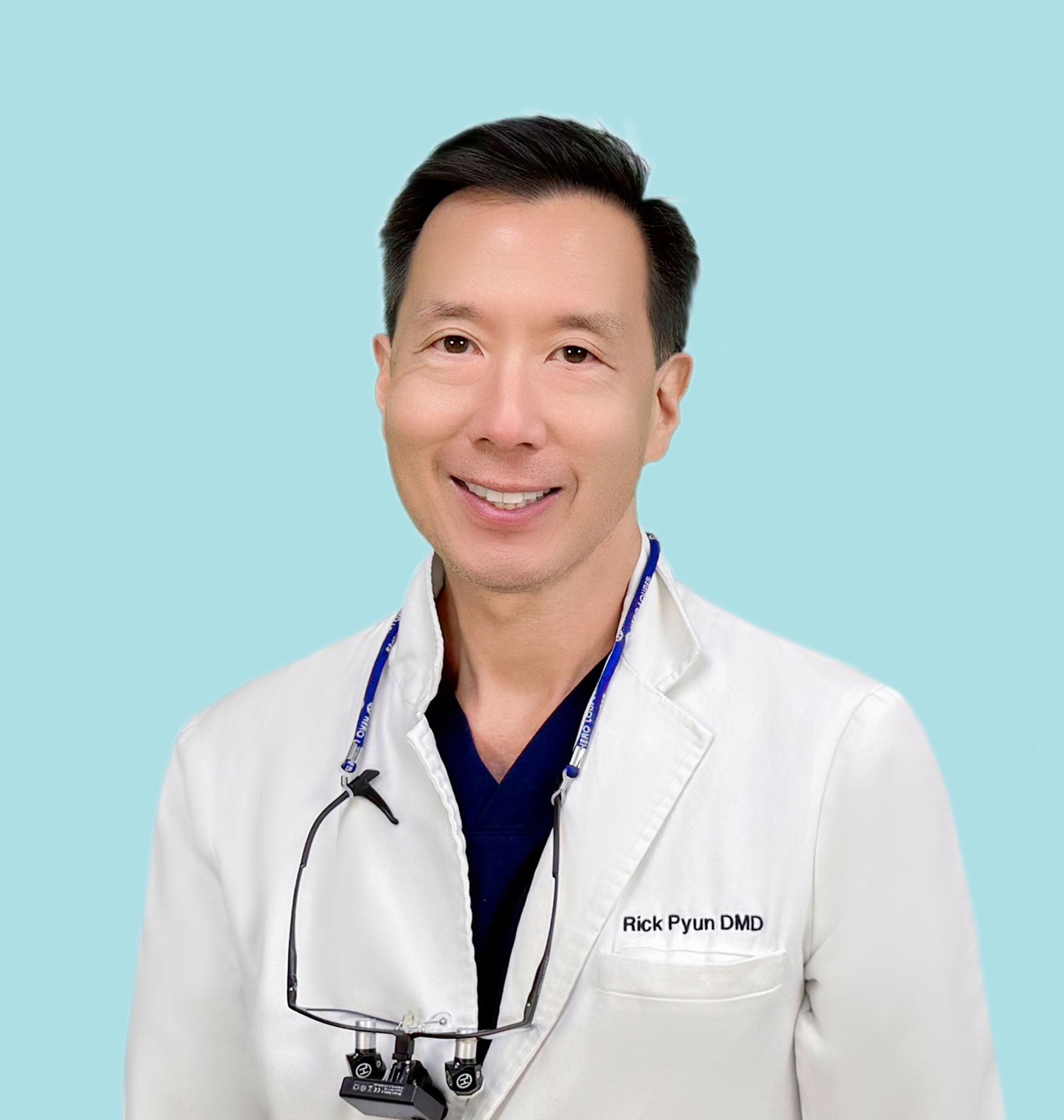 Smiling Dr. Ricky Pyun in a white lab coat, dental loupes, on a turquoise background. Rick Pyun, DMD, is on his coat.