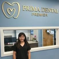 Woman standing in front of Prima Dental Premier sign. She wears a black top and smiles.