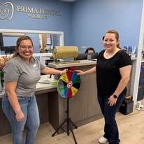 Two women at a Prima Dental office with a prize wheel. One spins, smiles; the other is smiling and has a prize.