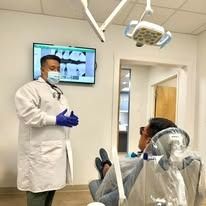 A dentist in a lab coat and mask explaining something to a patient in a dental chair.