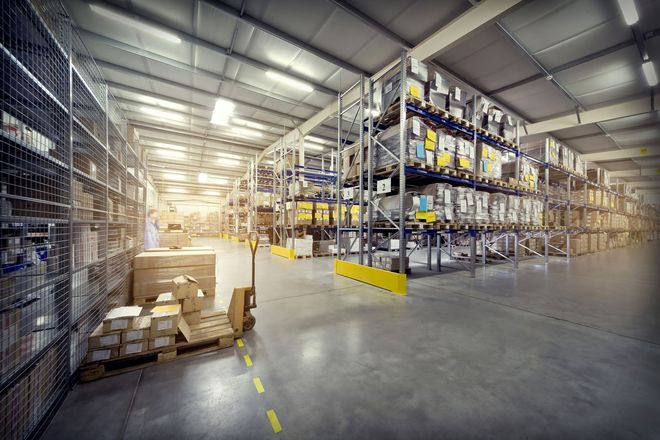 Wide warehouse aisle with tall metal racks, stacked boxes, and bright overhead lights