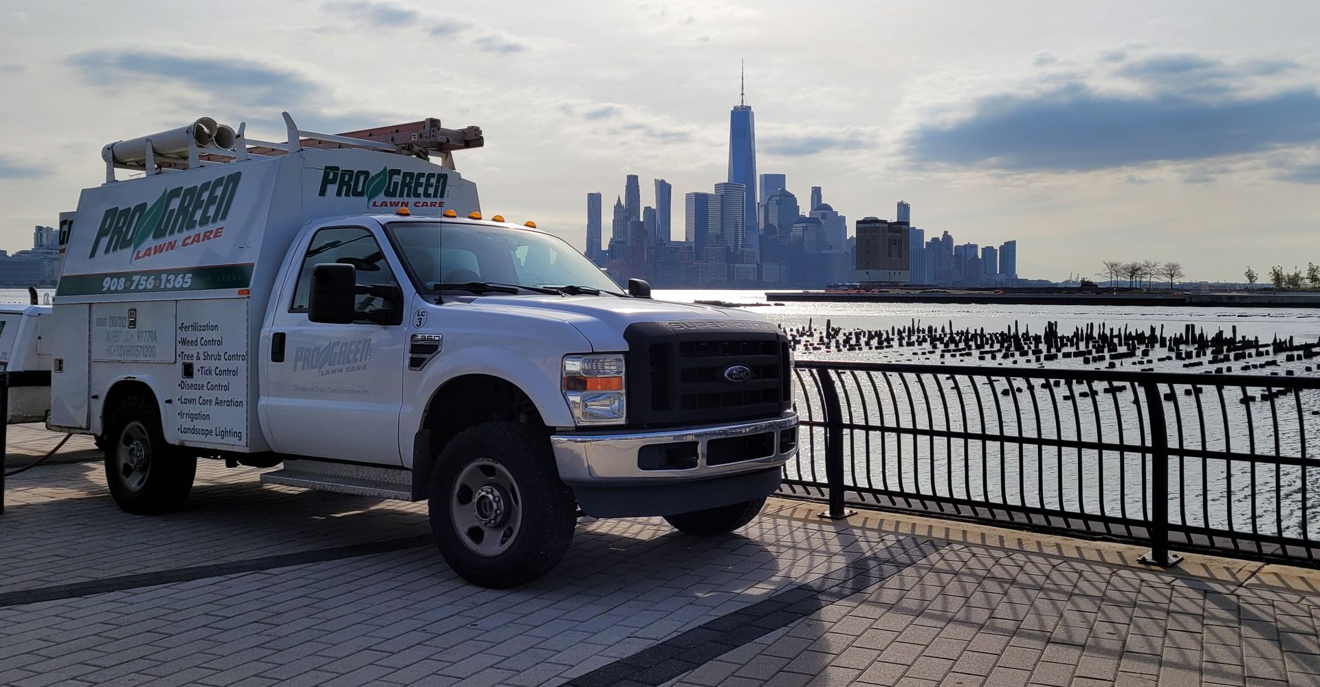 A white truck is parked next to a fence with a city skyline in the background.