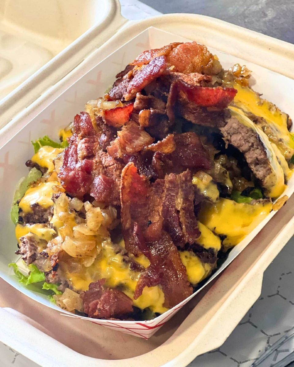 A takeout container filled with a burger bowl featuring chopped beef patties, melted cheese, bacon, and greens.