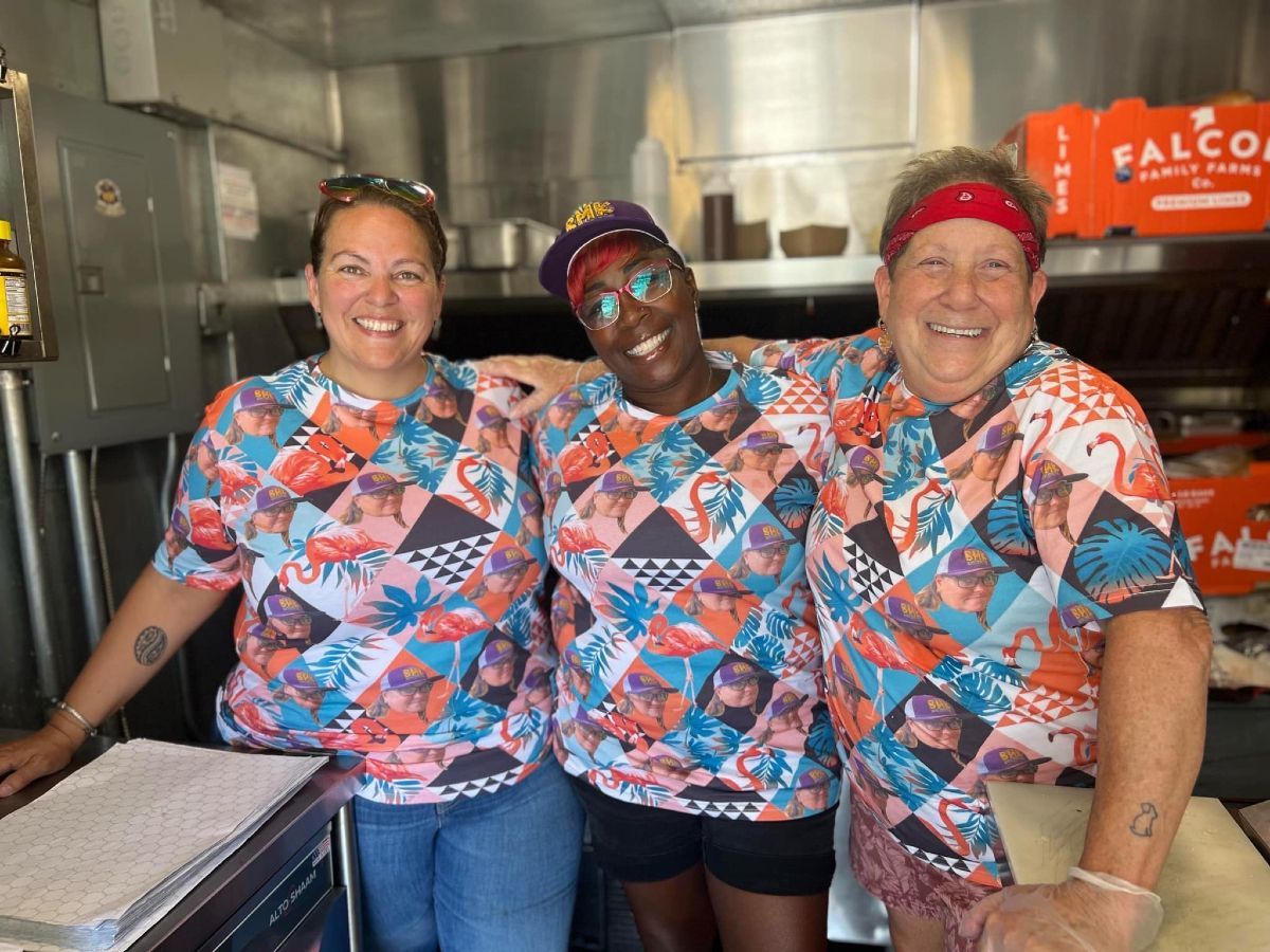 Three smiling coworkers wearing matching colorful patterned shirts and headbands stand side-by-side in a commercial kitchen.