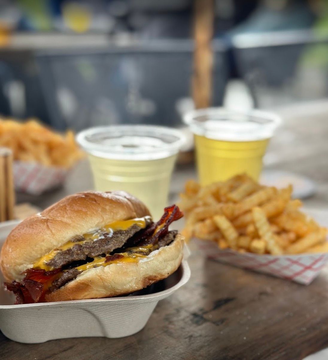 A bacon cheeseburger in a paper container next to crinkle-cut fries and two cups of light-colored drinks on a table.