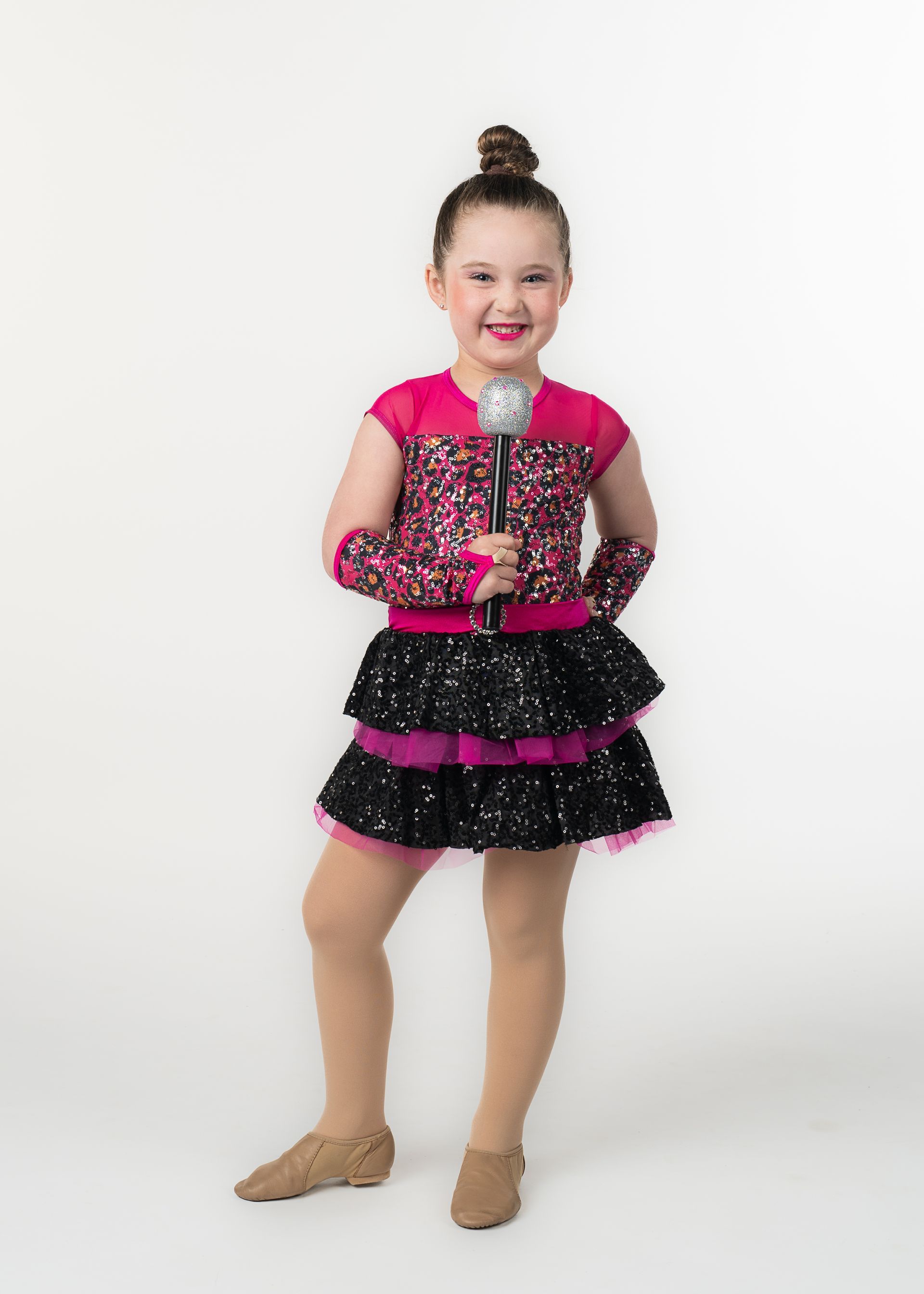 Young girl in a sequined dance costume holds a microphone and smiles.