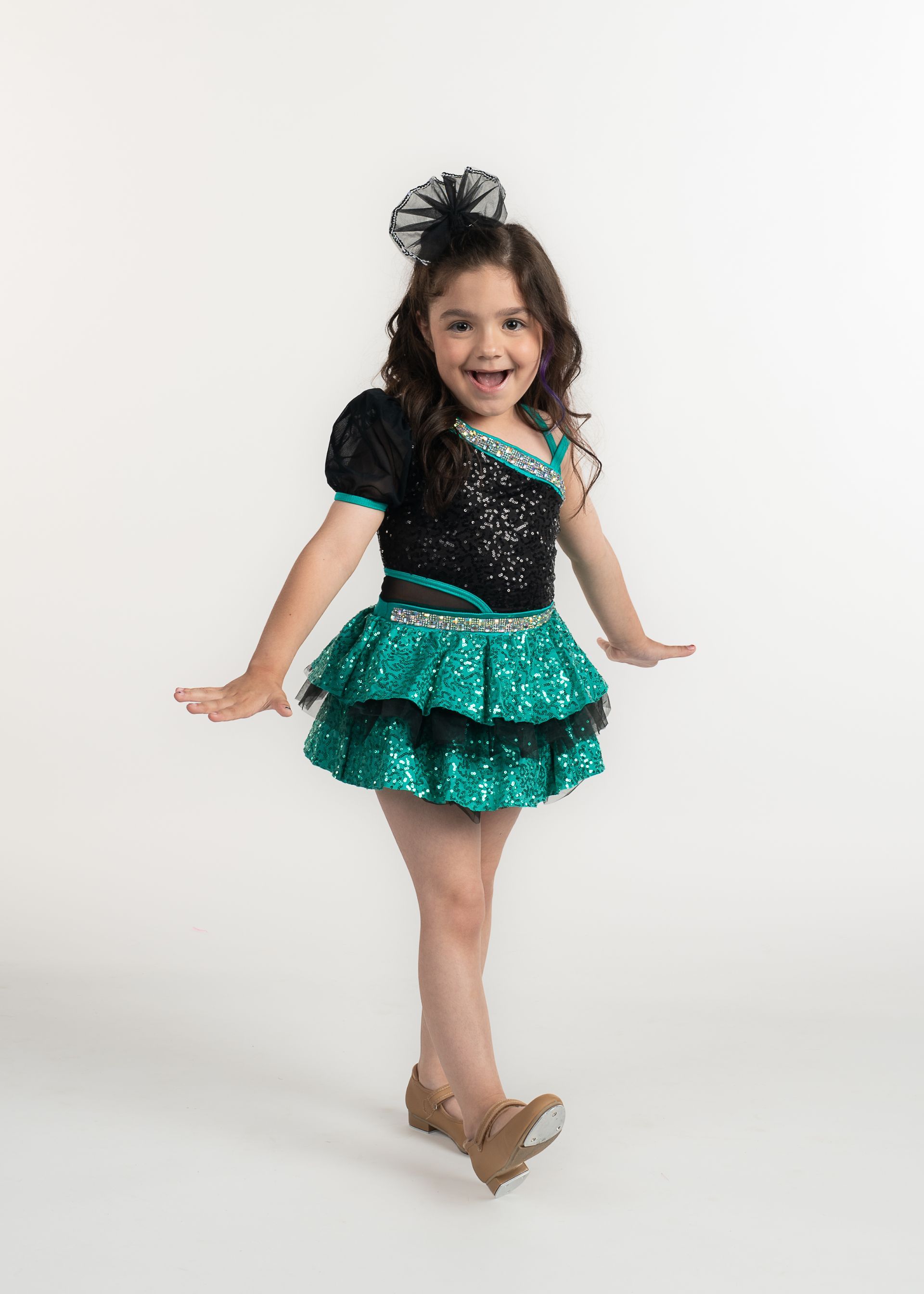 Girl in a dance costume, smiling, poses for the camera. Black and teal outfit, stage setting.
