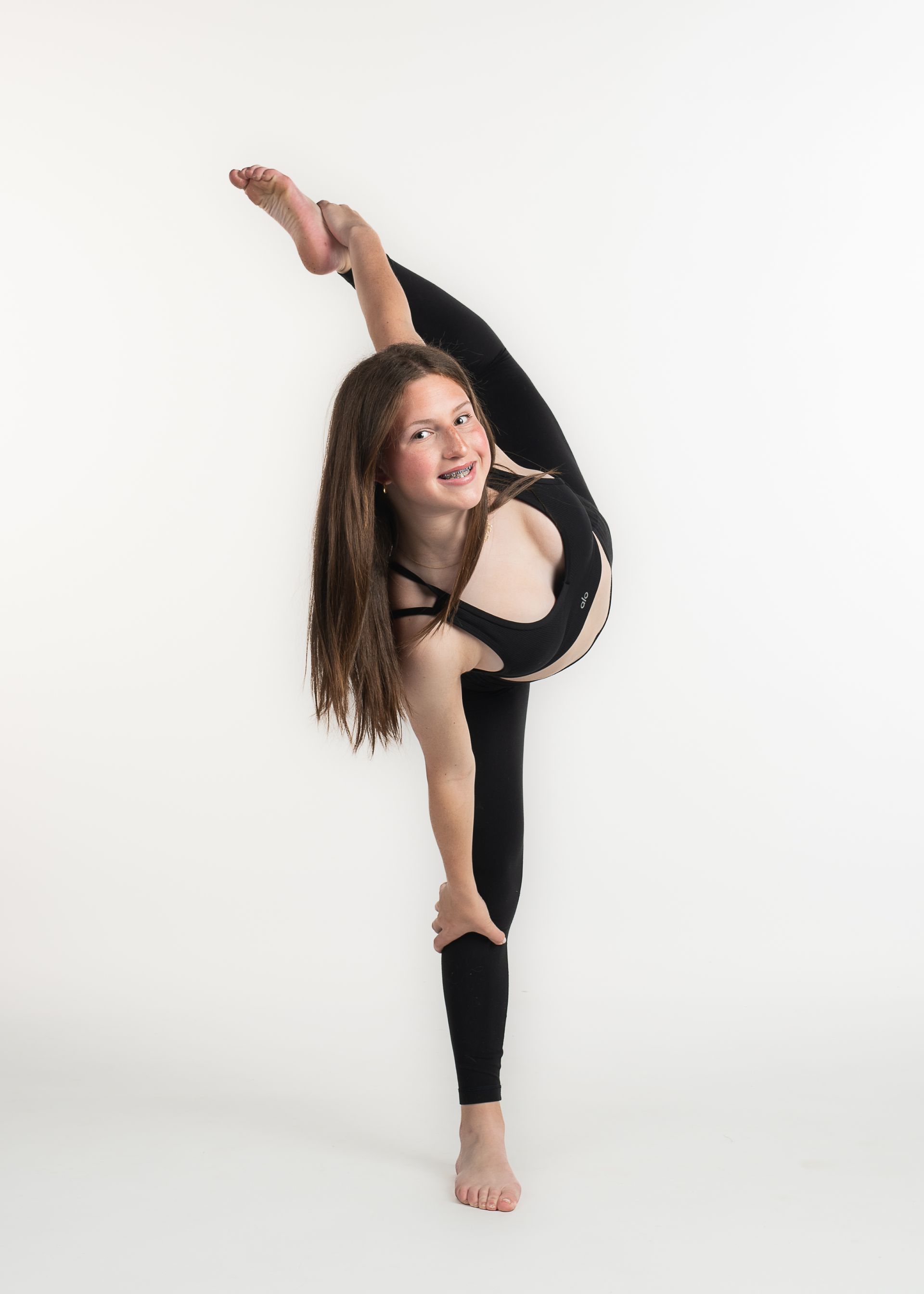 Teen in black activewear does a split with one leg raised, smiling against a white backdrop.