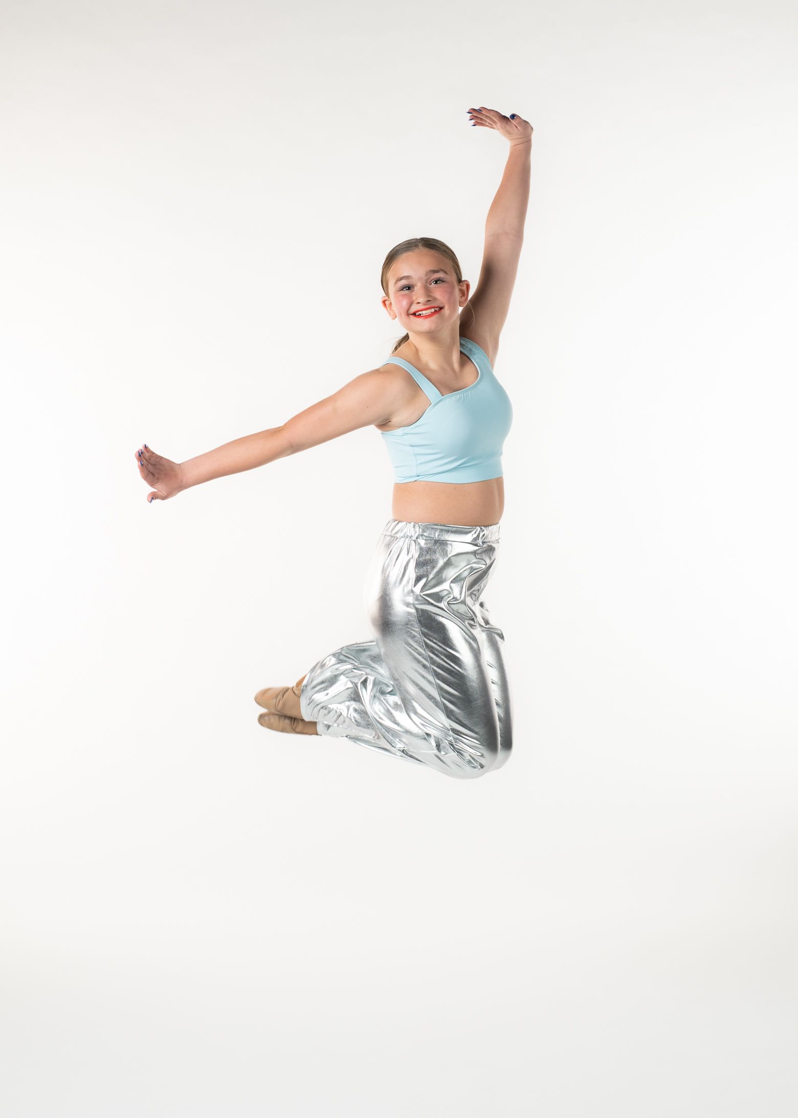 Dancer in silver pants and blue top, jumping with arms outstretched against a white background.