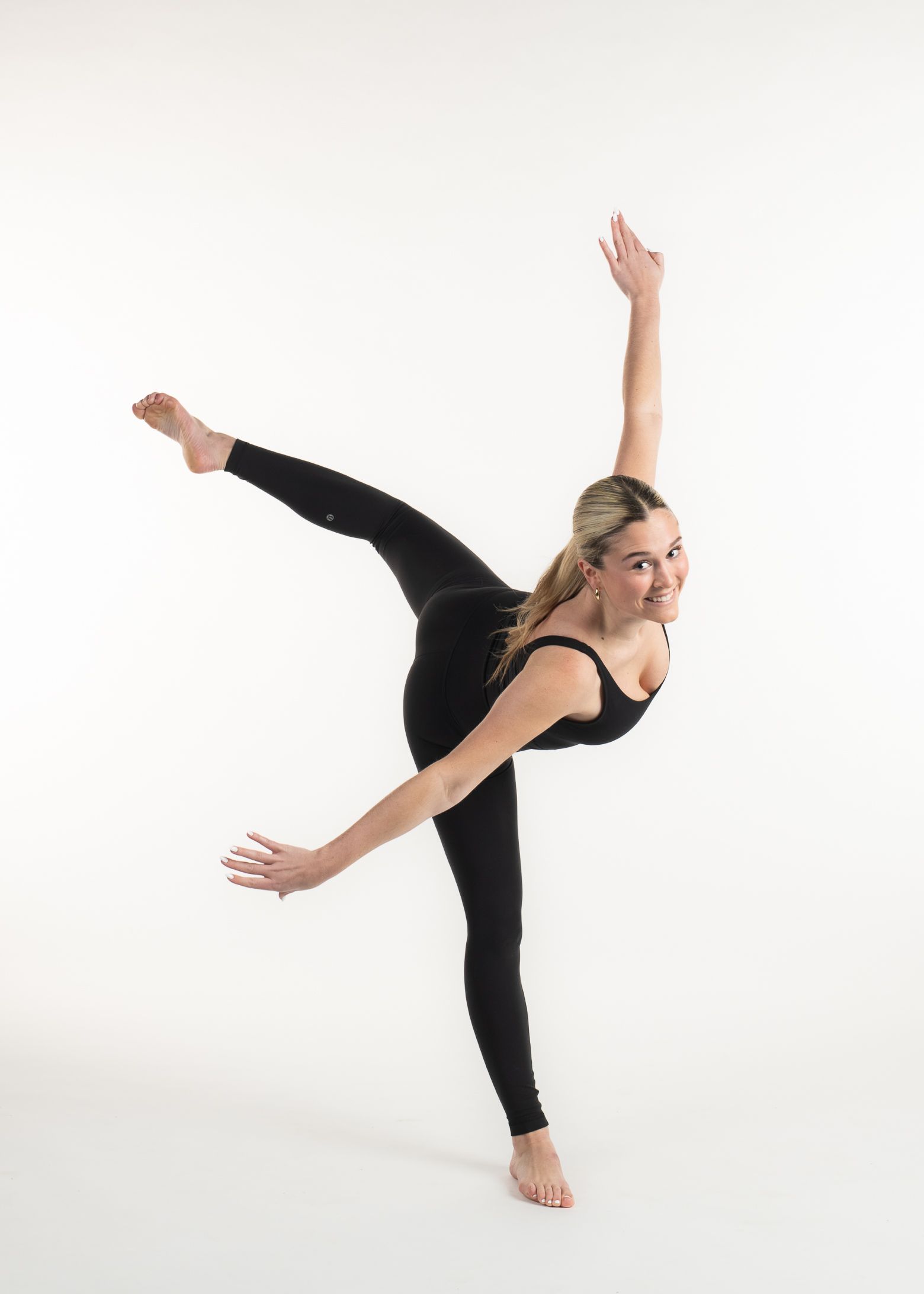 A young woman in black dance attire performs a graceful pose with a smile against a white backdrop.
