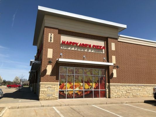 Exterior of Happy Joe's Pizza restaurant with brick and stone facade, large windows, and a clear blue sky.