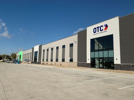 Exterior of an OTC building with a blue sky. Gray and white facade, glass entrance, and a parking lot.