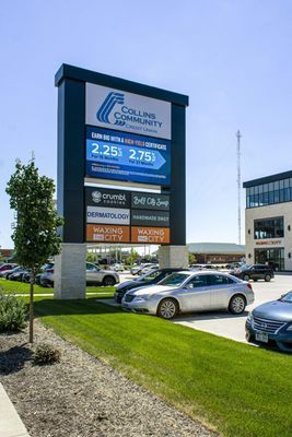 Sign for Collins Community, showing prices, with various store logos. Cars parked in the lot on a sunny day.