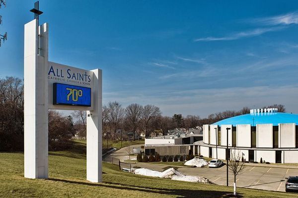 Sign for All Saints Church with temperature display and blue-roofed building on a sunny day.