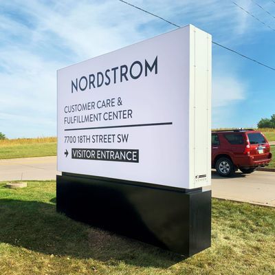 Sign for Nordstrom Customer Care & Fulfillment Center. White sign with black text, visitor entrance arrow. Black base. Red SUV parked.