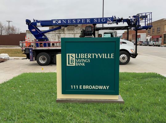 Sign for Libertyville Savings Bank at 111 E. Broadway; green sign with white text, installed by a truck with a lift.