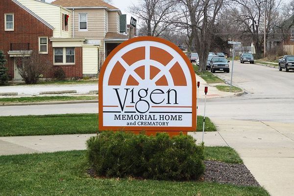 Sign for Vigen Memorial Home: Orange and white sign with arched top, text, and decorative design, near street and buildings.