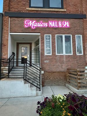 Exterior of Marion Nail & Spa with a pink neon sign over the entrance, brick building, and a small flower bed.