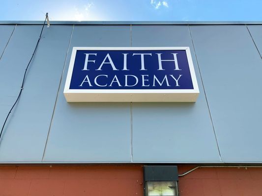 Sign for Faith Academy, blue and white text on a building, under a blue sky.