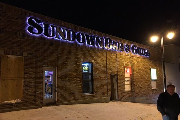 Sundown Bar & Grill at night, lit signage on brick building; snowy ground, person outside.