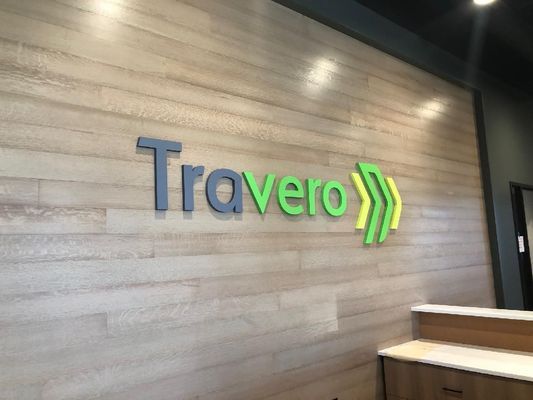 Travero logo on a light wood grain wall, with gray 