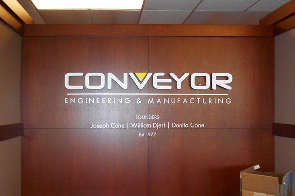 Signage for Conveyor Engineering & Manufacturing on a wood-paneled wall. Company name is white, and the logo includes a yellow triangle.