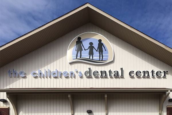 Sign for The Children's Dental Center. It features a graphic of three figures, with silver text on a beige building.