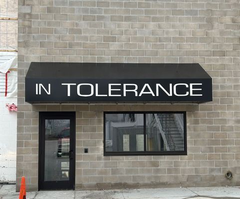 A storefront with a black awning that reads IN TOLERANCE over a door and a window.