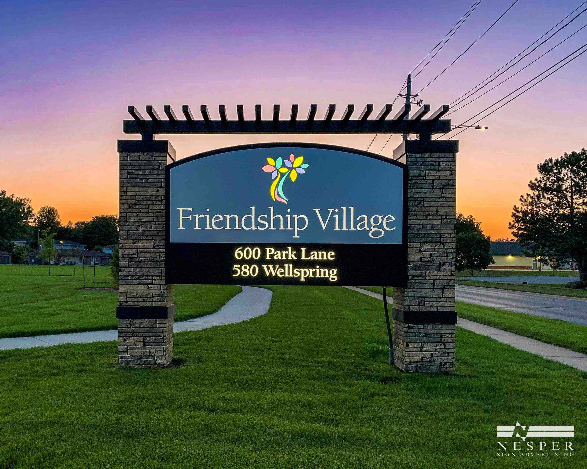 Friendship Village sign with colorful logo, address, and stone columns against an evening sky.