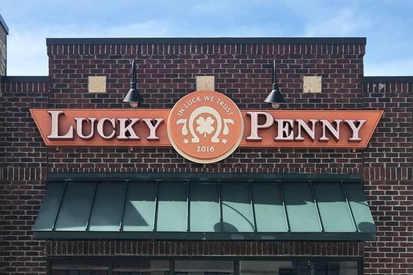 Lucky Penny restaurant sign on a brick building, with orange lettering and a circular logo featuring a horseshoe and shamrock.