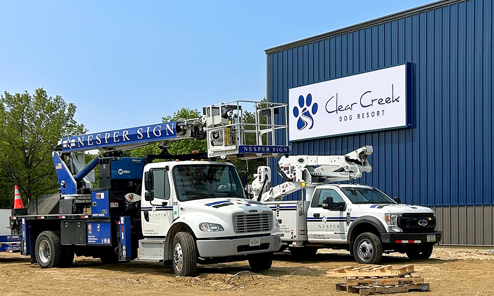 Two Clear Creek power trucks parked outside a blue building with sign; one truck has a raised boom.