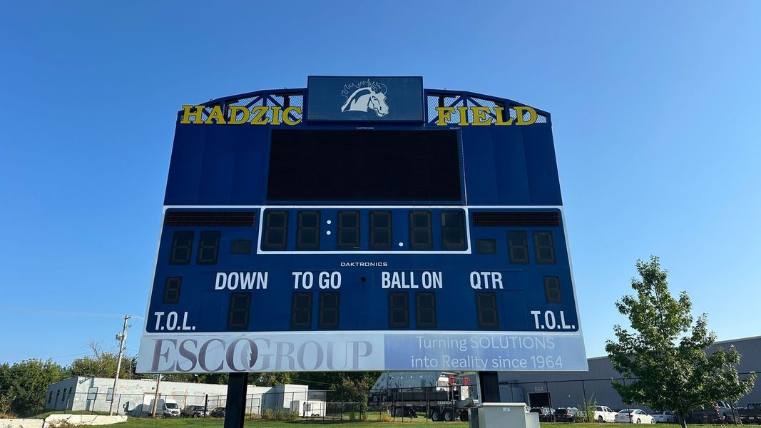 Blue and white football scoreboard under a clear sky; sponsored by ESCO Group.