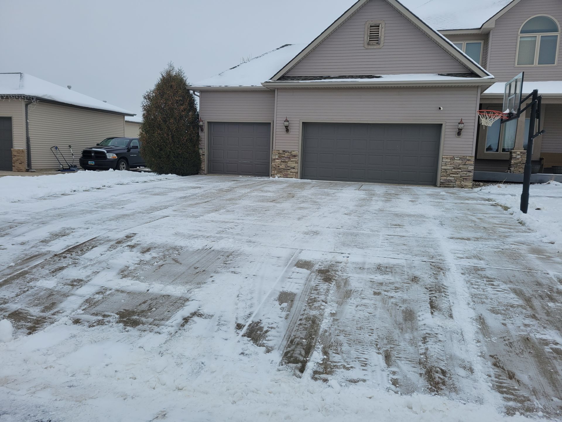 Snow-covered driveway in front of a house with two garage doors. A basketball hoop is visible.