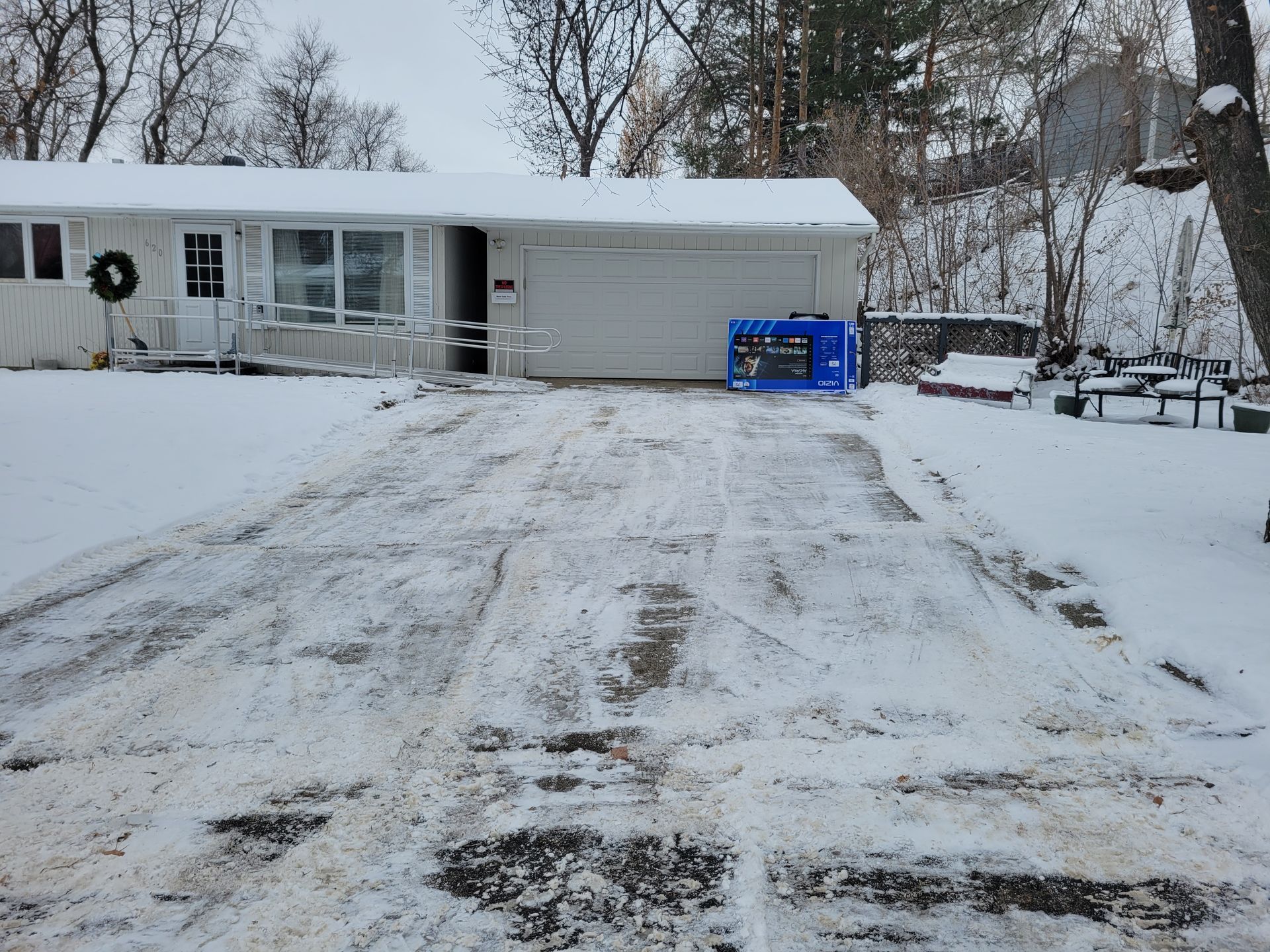 Snow-covered driveway leads to a house with a garage; a generator sits in front. Trees and snow in background.
