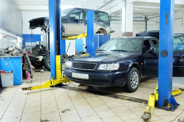 Cars in a brightly lit auto repair shop lift area, including a black sedan in front.