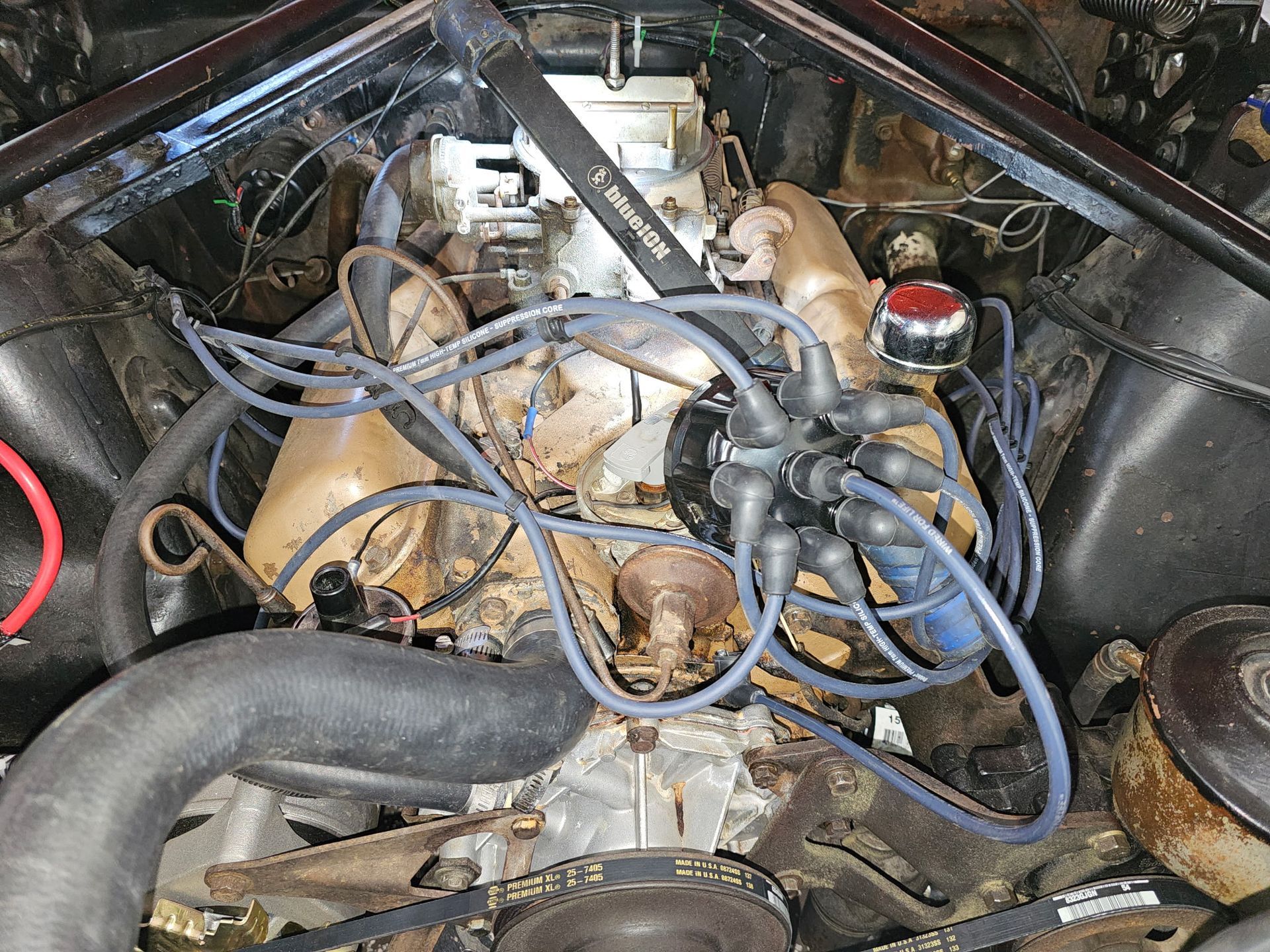 Car engine bay with blue wires, hoses, and rusted metal parts under the hood