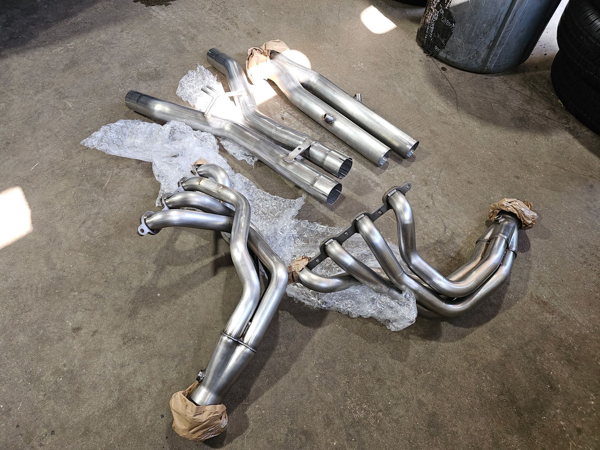 Several shiny metal exhaust headers and pipes laid on a carpeted floor, wrapped in bubble wrap.