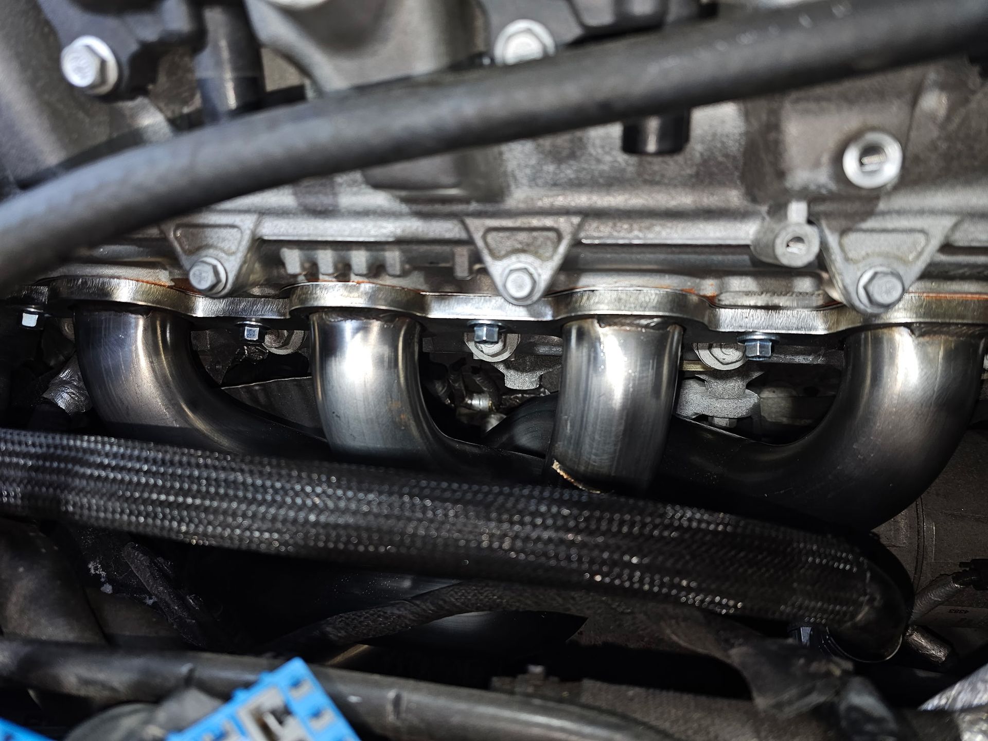 Close-up of shiny metal exhaust headers and engine components with black braided hose in the foreground