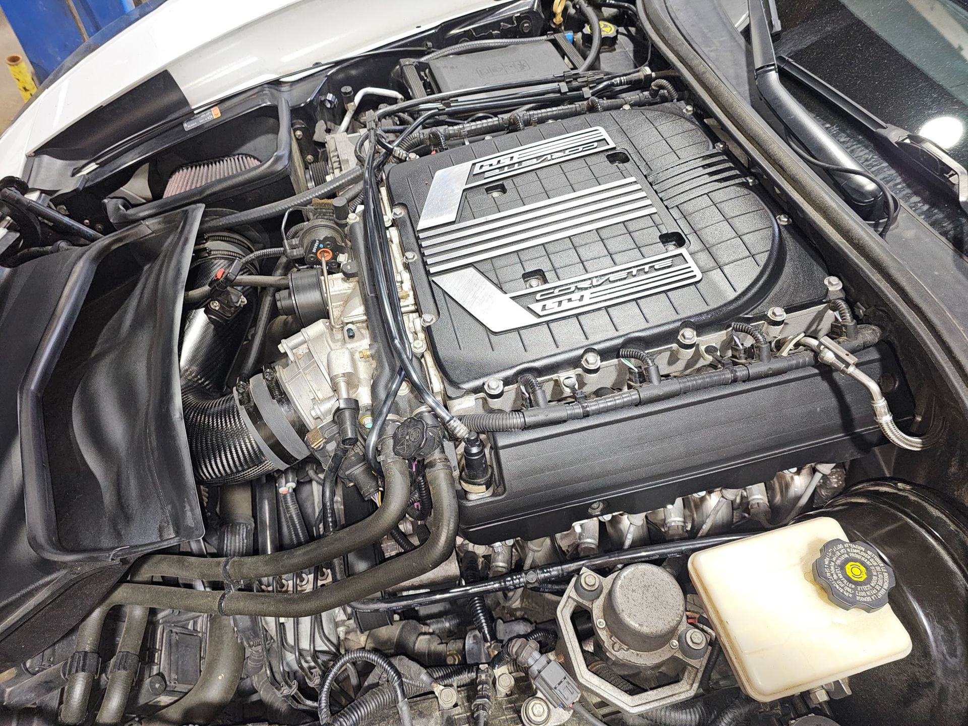 Car engine bay with a large air intake cover and visible hoses, battery, and coolant reservoir.