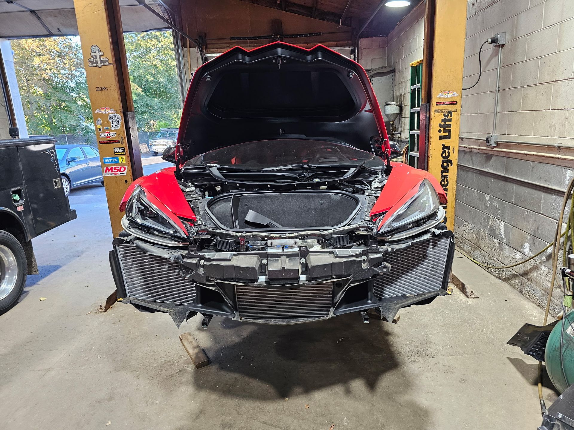 Damaged red car with raised hood in a garage, front bumper torn off and engine exposed