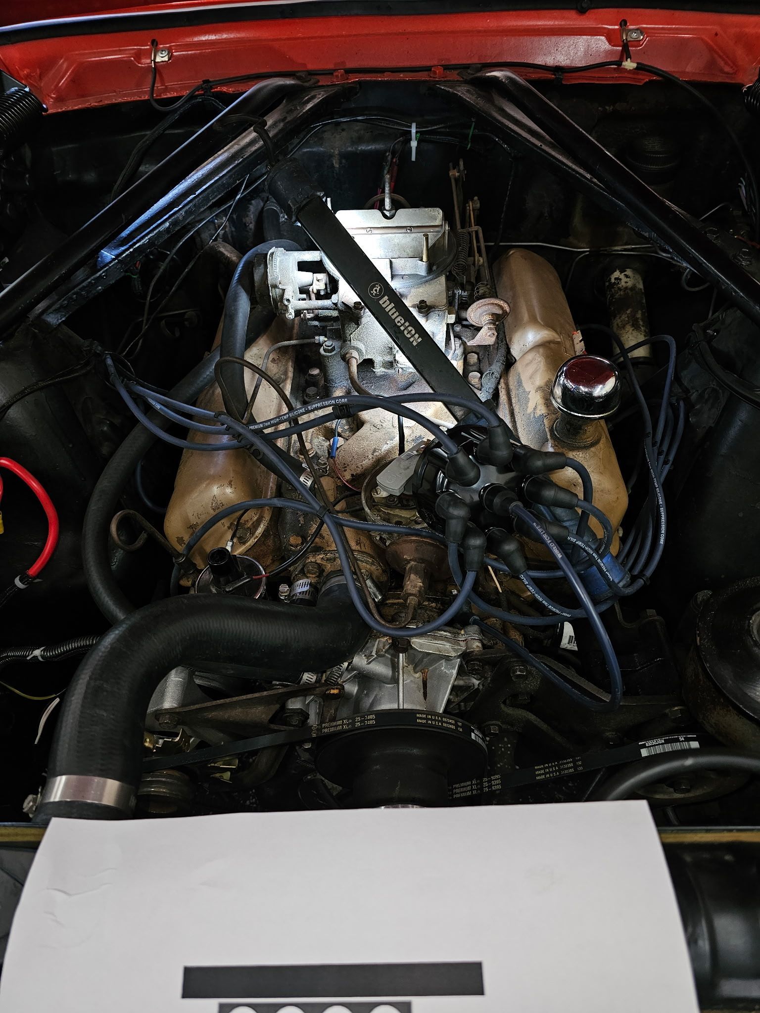 Open car engine bay with exposed components and tangled black hoses, red hood framing the top.