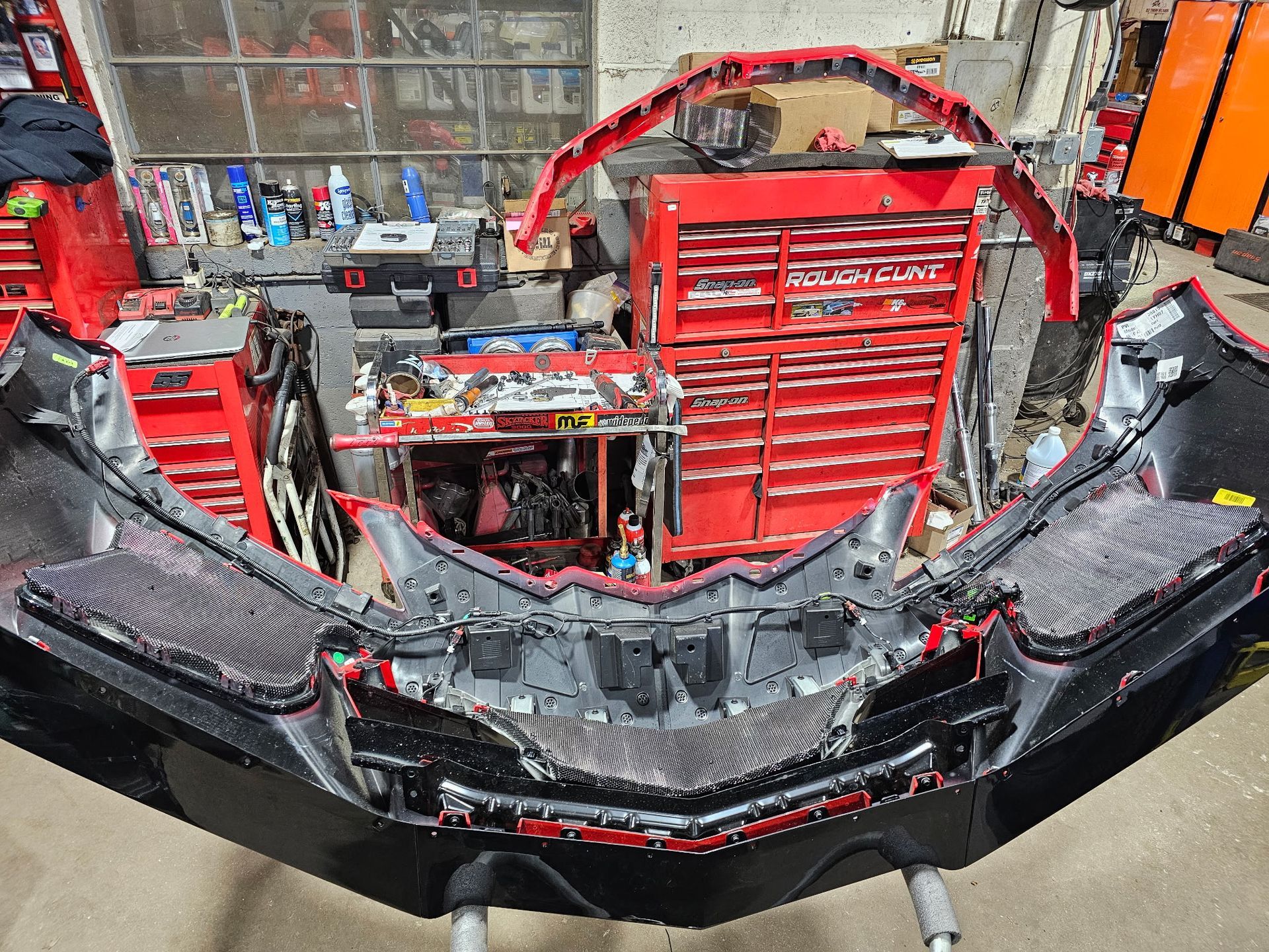 Car front bumper removed in an auto repair shop, showing exposed interior and red tool cabinets behind it
