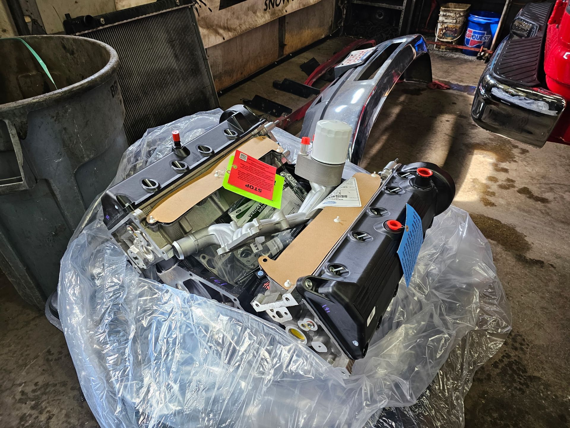A damaged car part wrapped in plastic sits on a wet garage floor with tools and containers nearby.